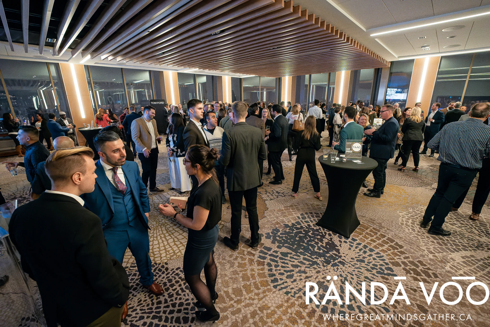 Guests in conversation around cocktail tables at a RÄNDĀ VO͞O reception beneath a wood-slat ceiling.