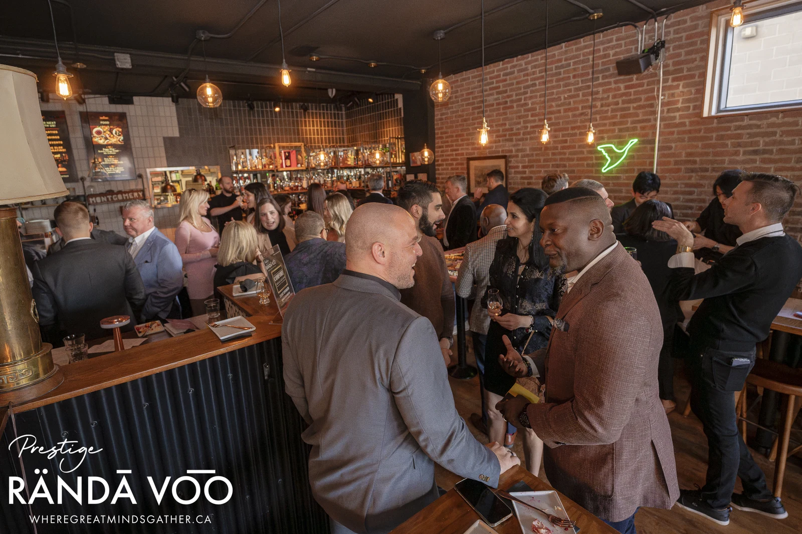 Guests sharing a handshake and conversation beside a brick-walled bar at a RÄNDĀ VO͞O Prestige gathering.