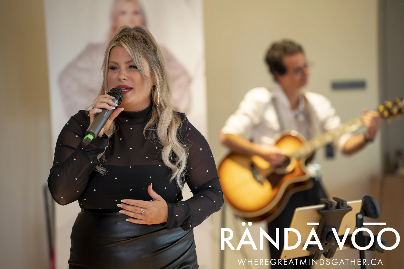 A vocalist singing into a microphone with a guitarist performing behind her at a RÄNDĀ VO͞O gathering.