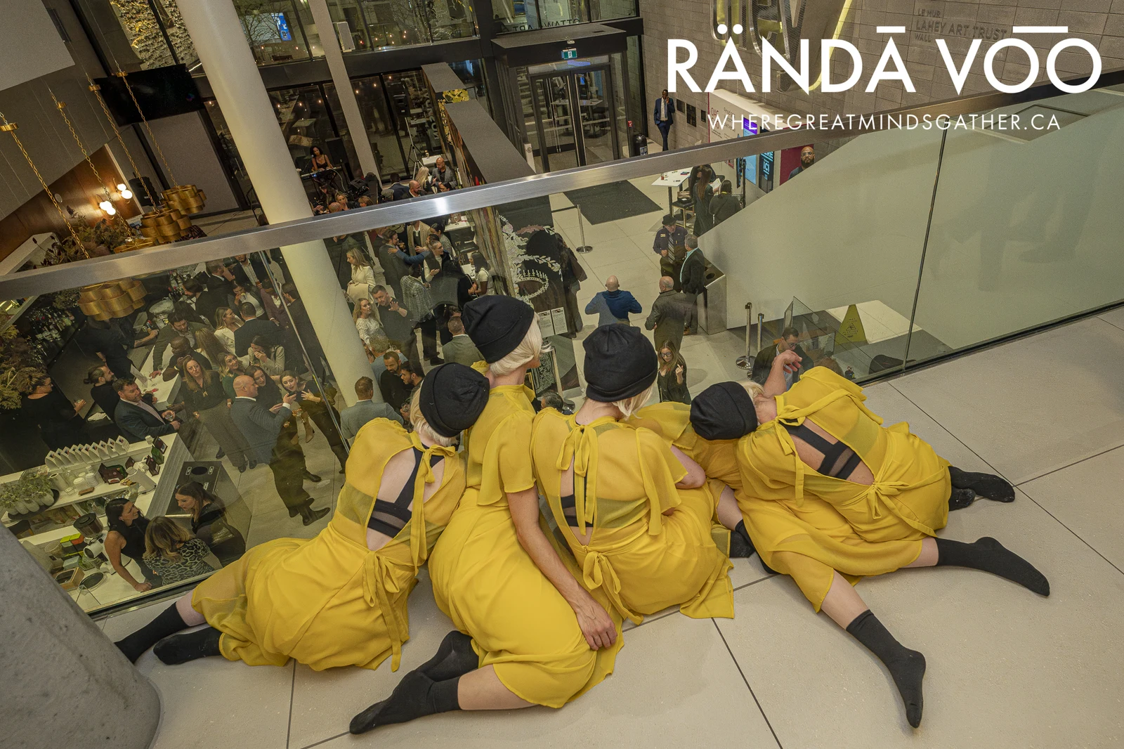 Four performers in yellow outfits seated above a reception, overlooking guests gathered in a multi-level venue.