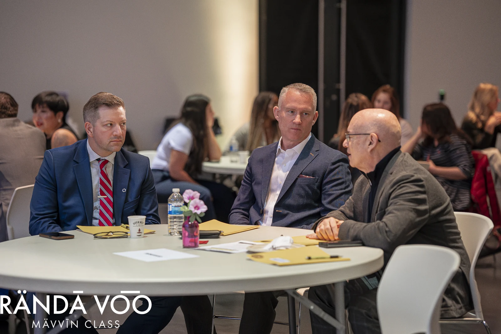 Three MÄVVĪNCLASS participants in conversation around a table with notebooks and refreshments.