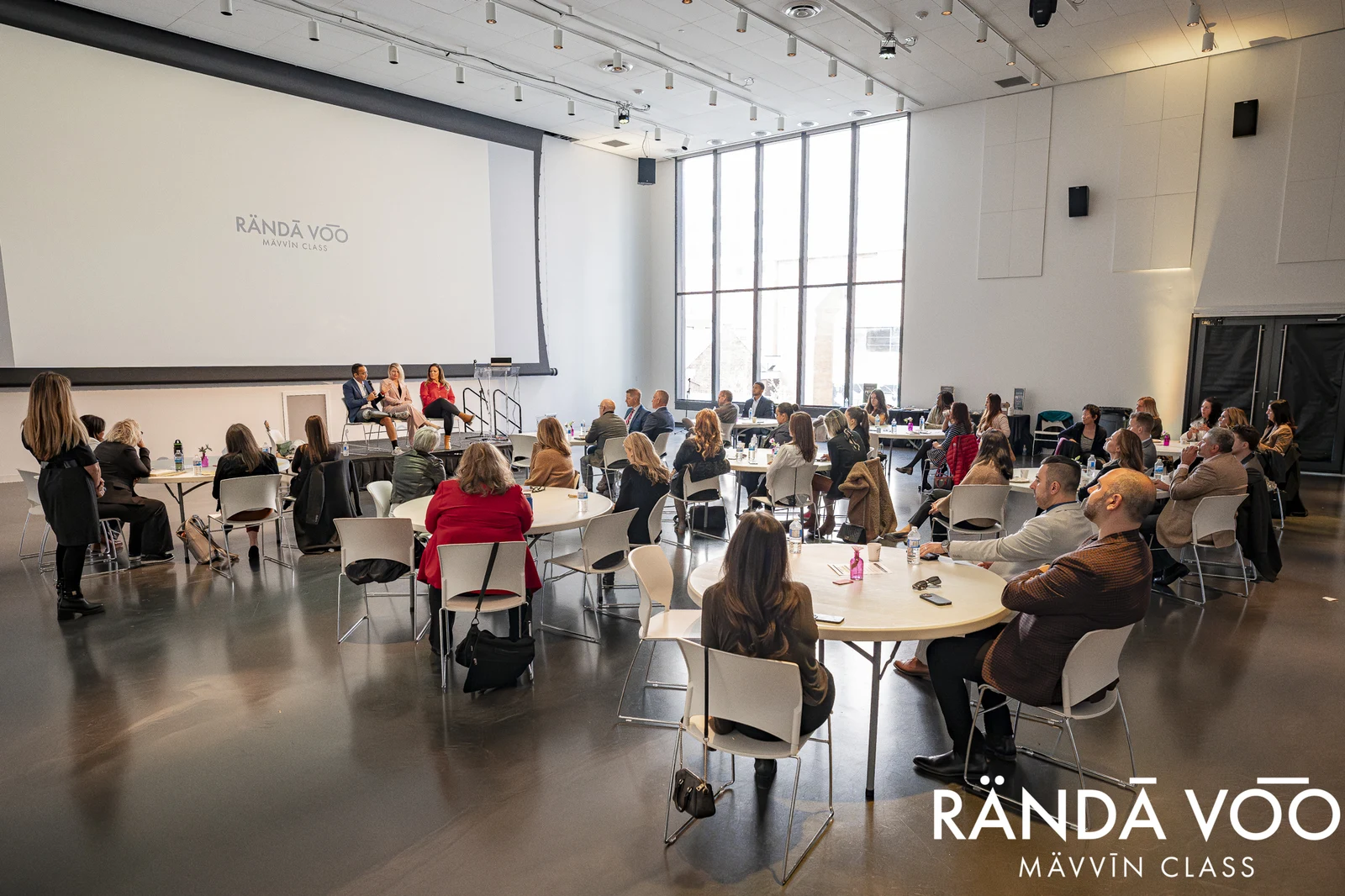 MÄVVĪNCLASS attendees seated around round tables facing a panel of three speakers in a bright presentation room.