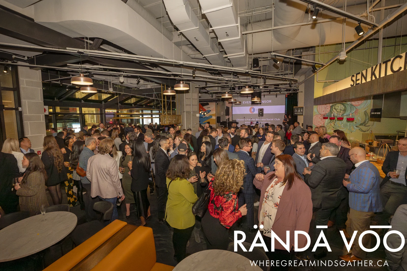 A busy RÄNDĀ VO͞O gathering with guests in conversation throughout a venue with exposed ceiling fixtures and food counters.