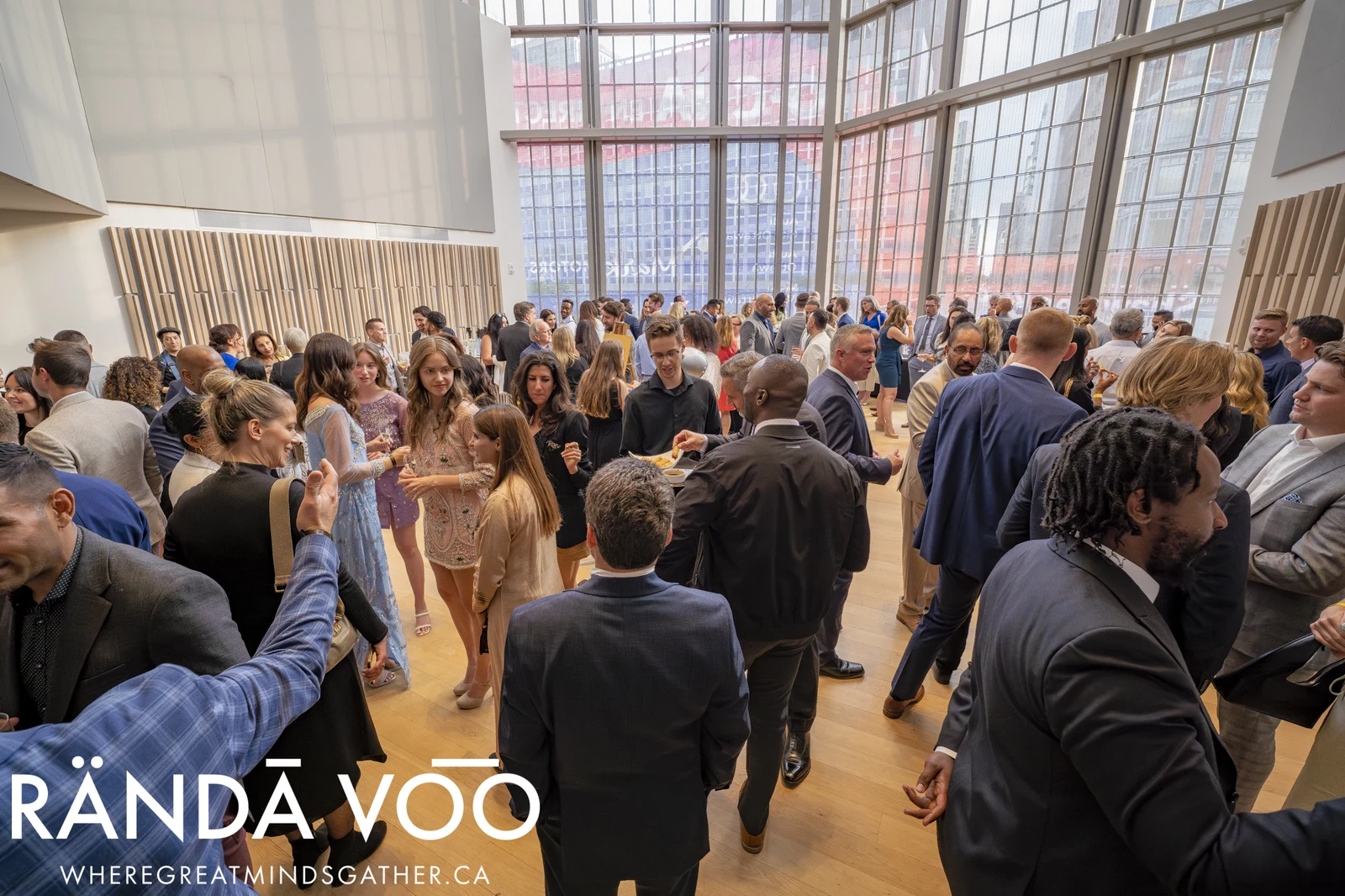 A large group of guests in conversation at a RÄNDĀ VO͞O reception in a bright venue with tall windows and wood-panelled walls.