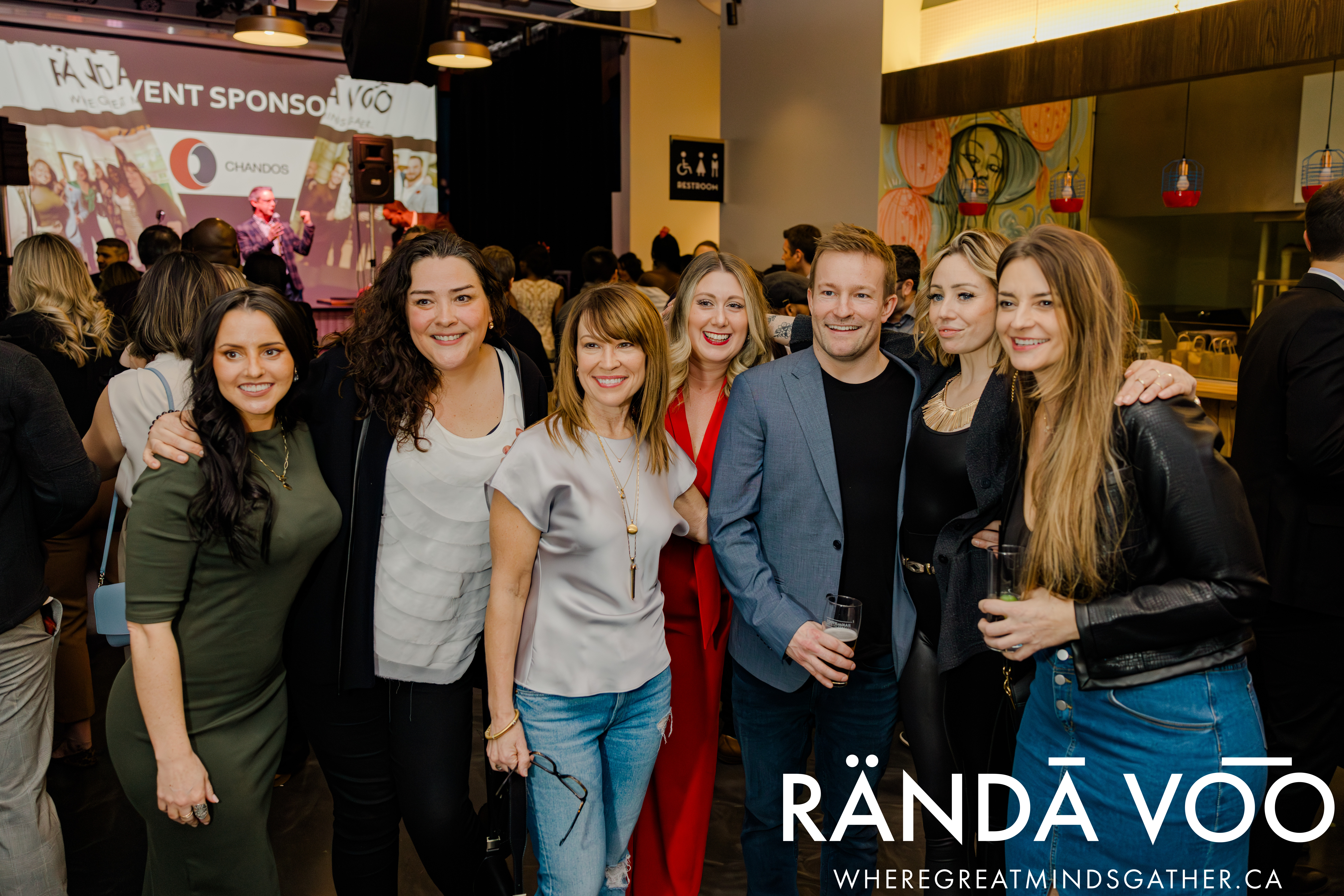 Guests posing together in front of the event stage at Queen St. Fare, with RÄNDĀ VO͞O branding.