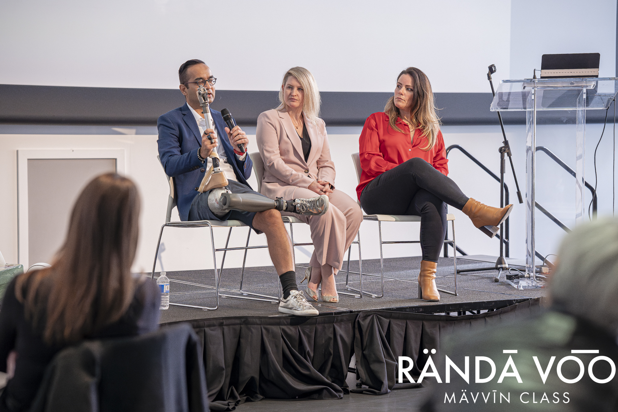 Three panelists on stage at a MÄVVĪNCLASS discussion for the RÄNDĀ VO͞O business community, with one speaking into a microphone.