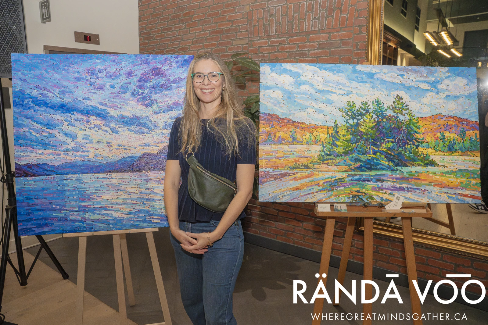 An attendee standing between two colourful landscape paintings displayed on easels at a RÄNDĀ VO͞O business community event.