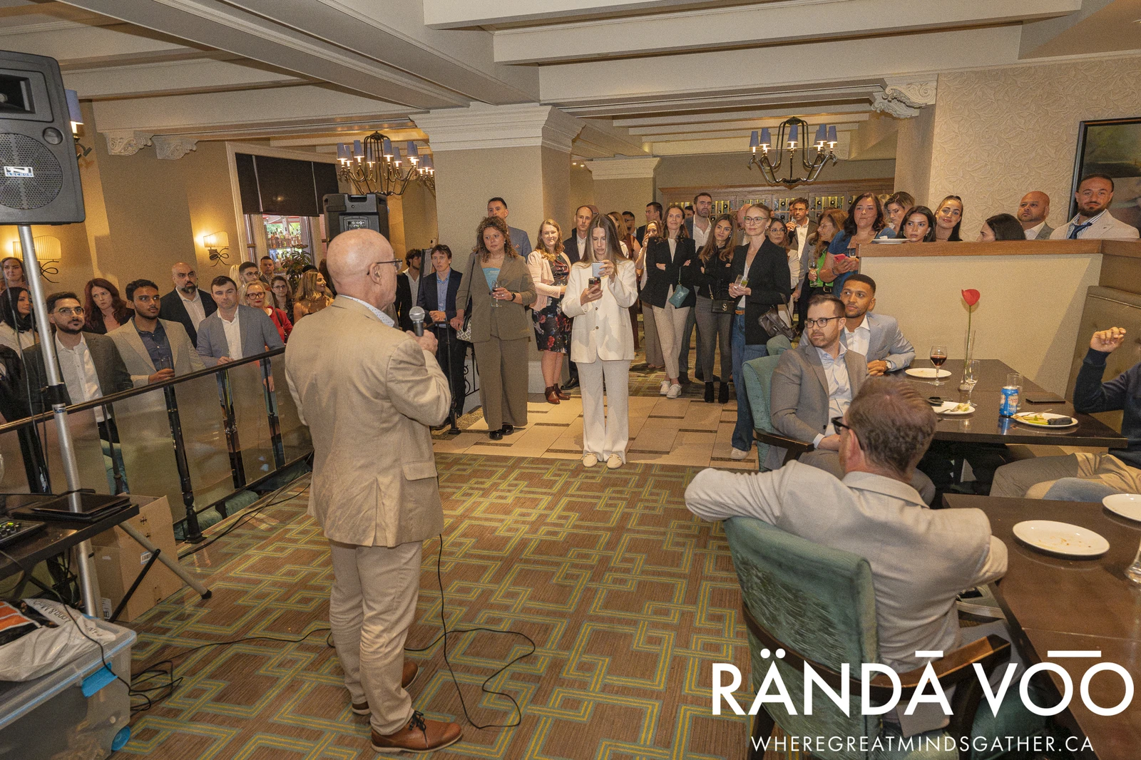 A speaker addressing RÄNDĀ VO͞O business community attendees gathered throughout a reception room.