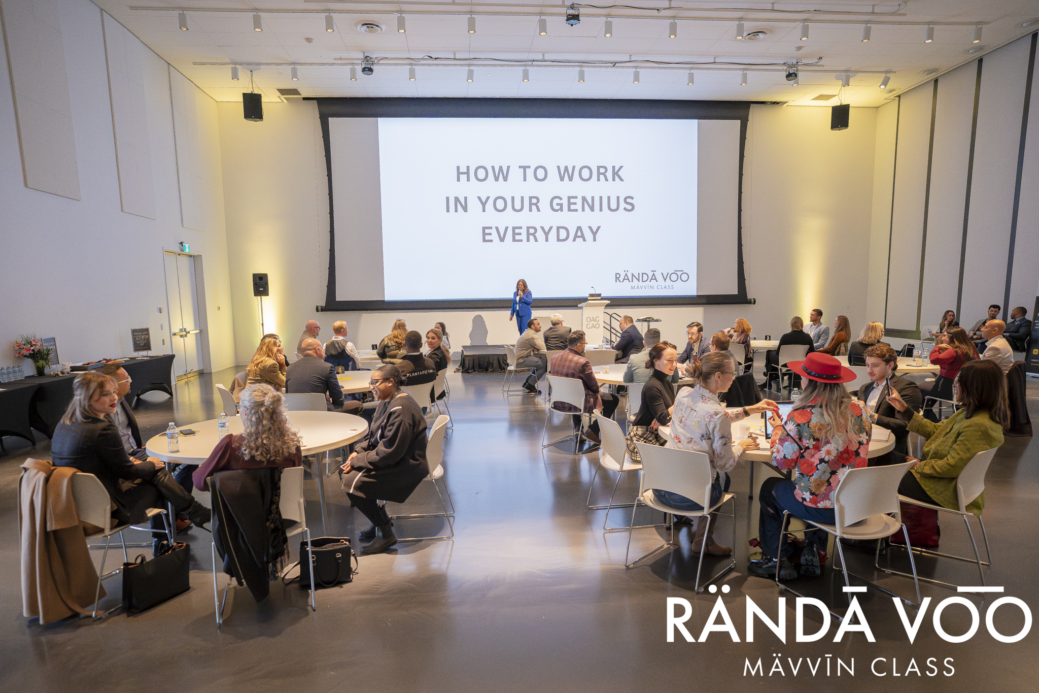 RÄNDĀ VO͞O business community members seated at round tables during a MÄVVĪNCLASS presentation, with a speaker beneath a screen reading “How to work in your genius everyday.”