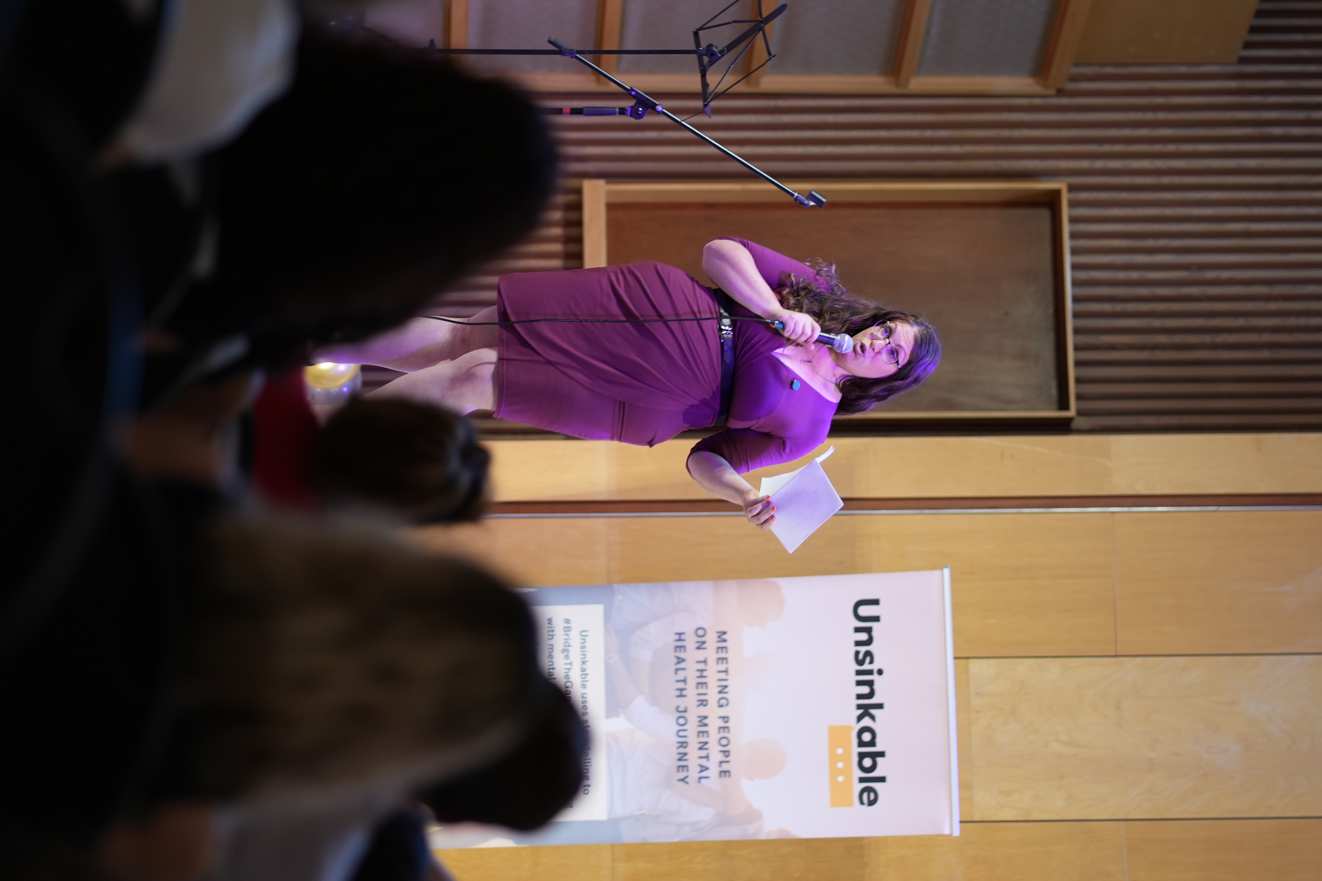 Hailey Hechtman speaking into a handheld microphone beside an Unsinkable banner, with audience members in the foreground.