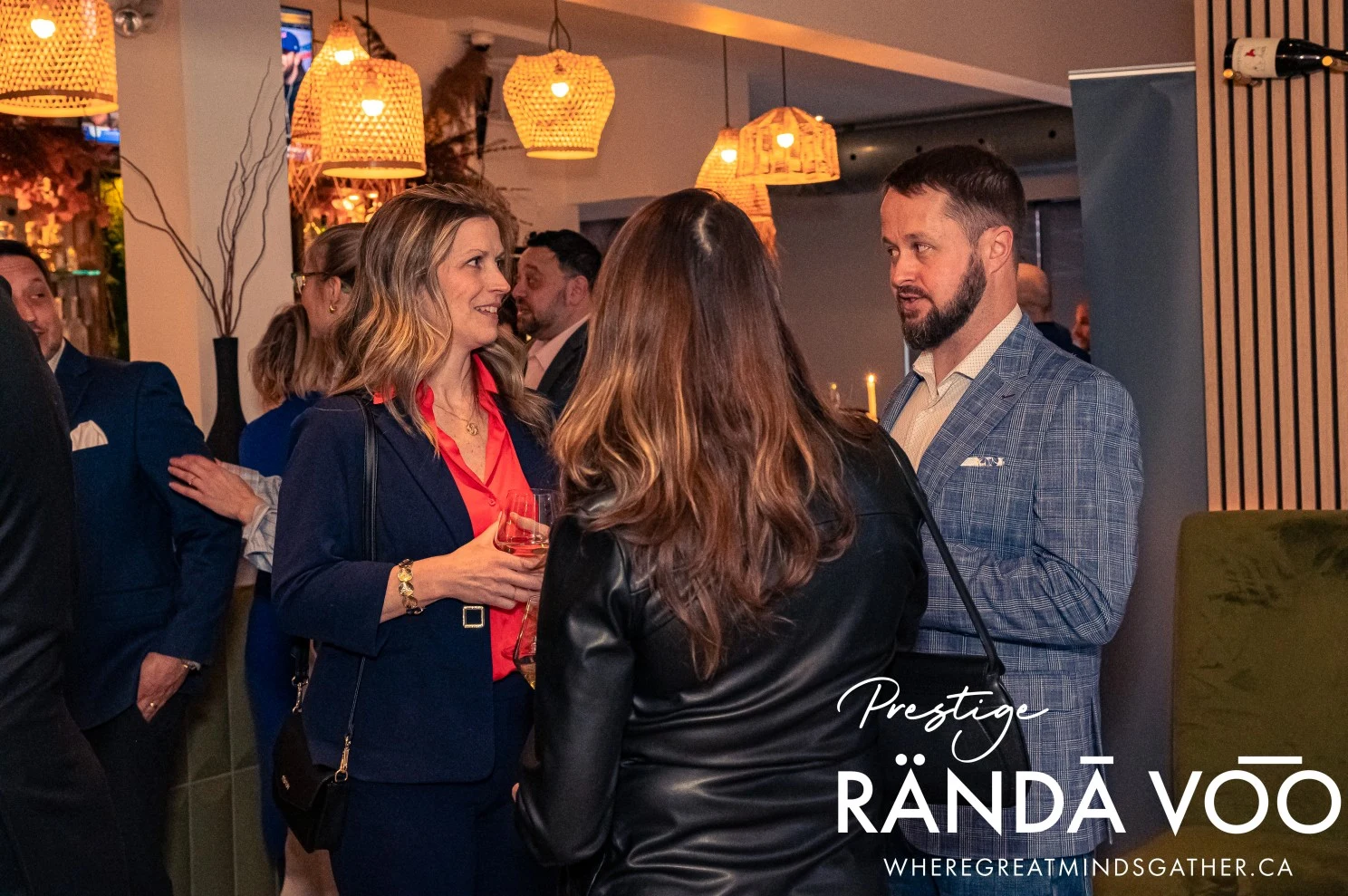 Guests in conversation beneath pendant lights at HEIST Manotick, with RÄNDĀ VO͞O Prestige branding.