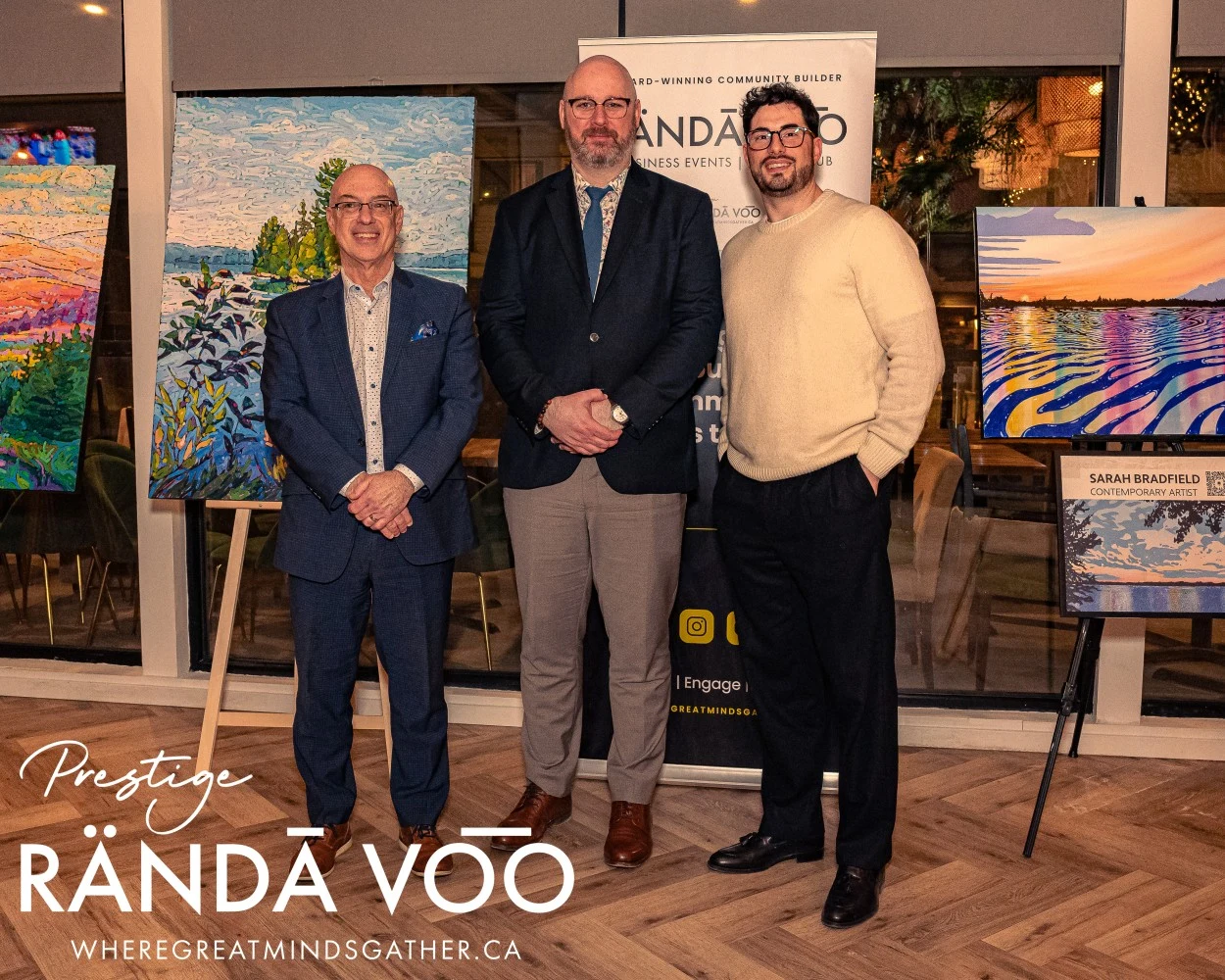 Three guests posing beside displayed paintings at HEIST Manotick, with RÄNDĀ VO͞O Prestige branding.