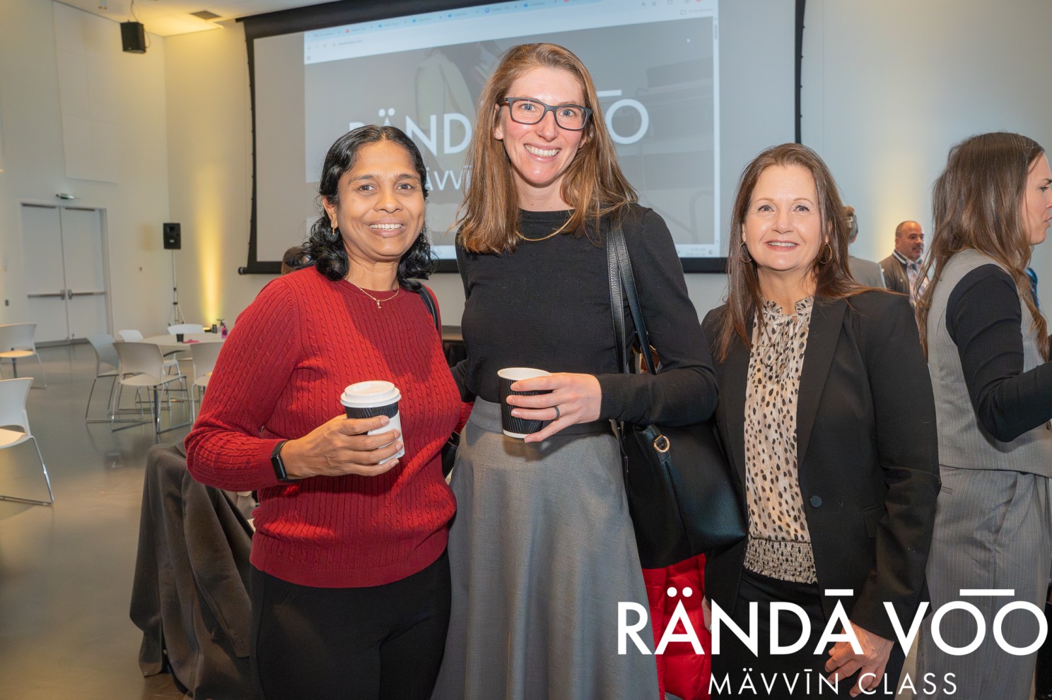 Three MÄVVĪNCLASS guests posing together with coffee in front of the projection screen.