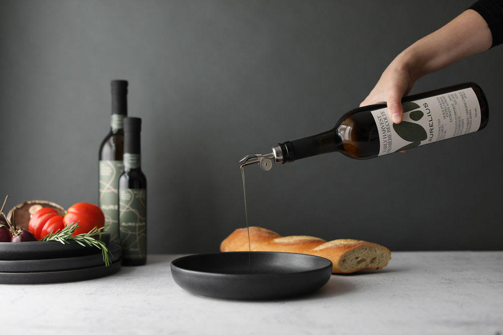 A hand pouring Aurelius olive oil into a black bowl beside bread and fresh produce.