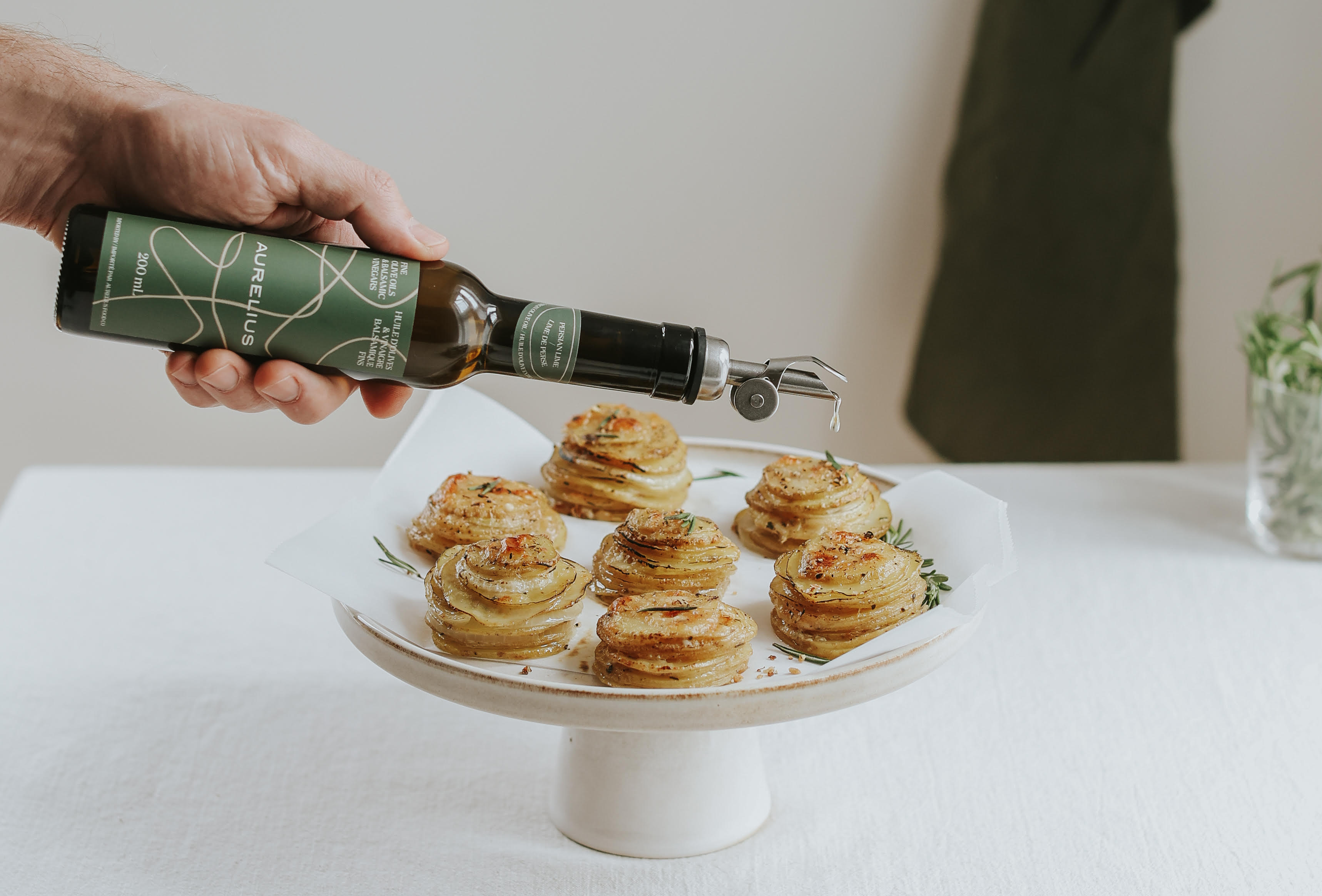 A hand pouring Aurelius olive oil from a bottle over prepared potato stacks on a serving stand.